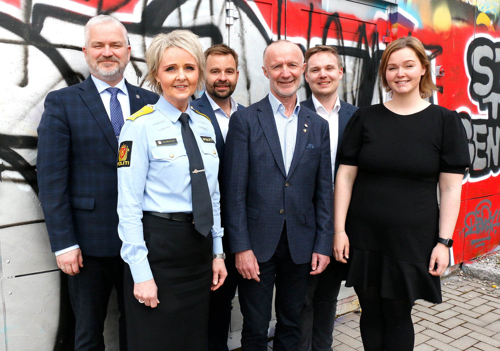 Fire menn i mørk dress, en kvinne med mørk kjole og en kvinne med blå skjorte og sort skjørt står utendørs foran en fargerik vegg.