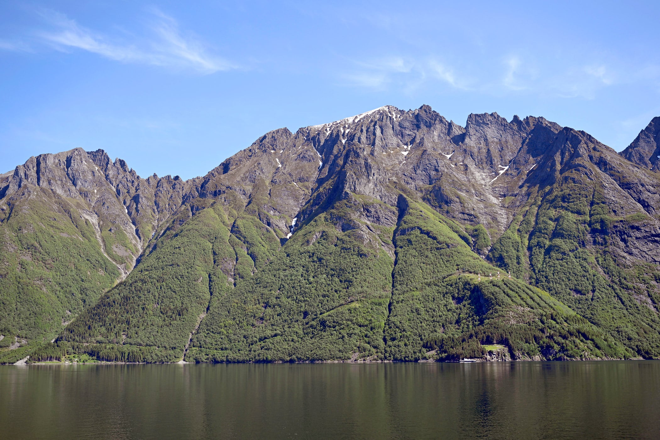Fjordlandskap med høge, bratte fjell dekte av grøn vegetasjon og nokre snøflekker nær toppane. Den stille fjorden i framgrunnen speglar fjella. Klar blå himmel med nokre få lette skyer.