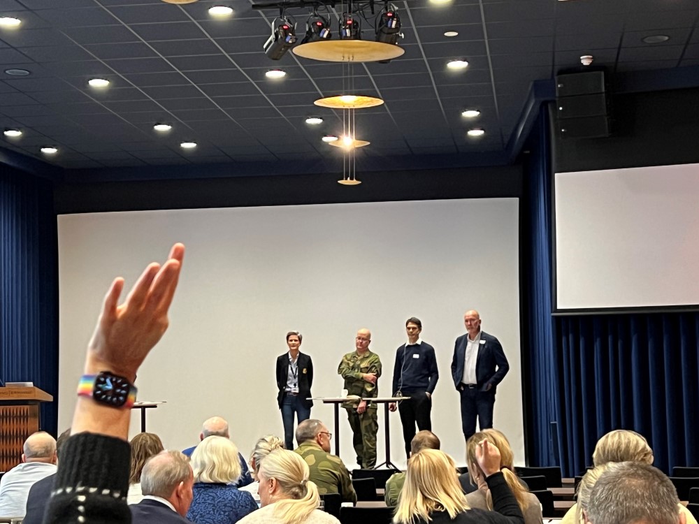 Menneske i auditorium med panel i front. Ein person rekker opp handa.