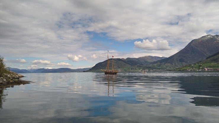 seglbåt utan segl, på fjord omkransa av høge fjell. Mindre båtar i følgje. 