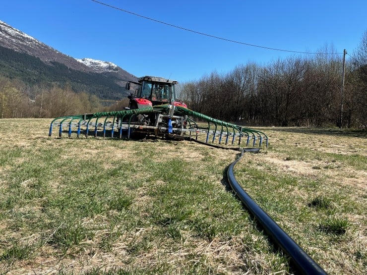 Blå himmel, fjell, skog,  rød traktor, gjødselspreiar