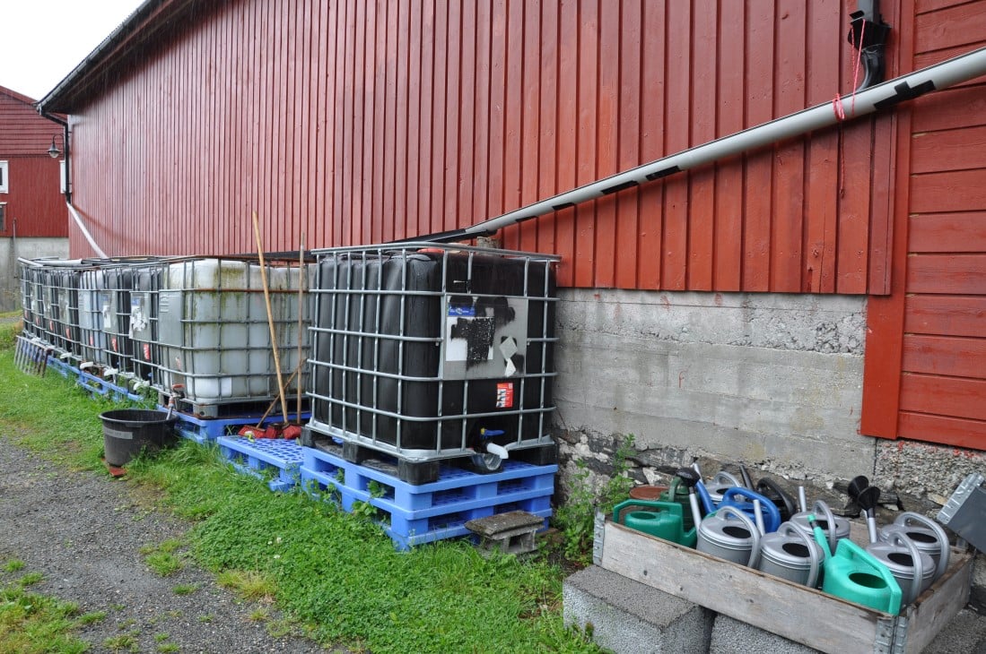Låvevegg med rør som leder ned vann i store 1000 liters kontainere. Vannkanner stablet ved siden av. 