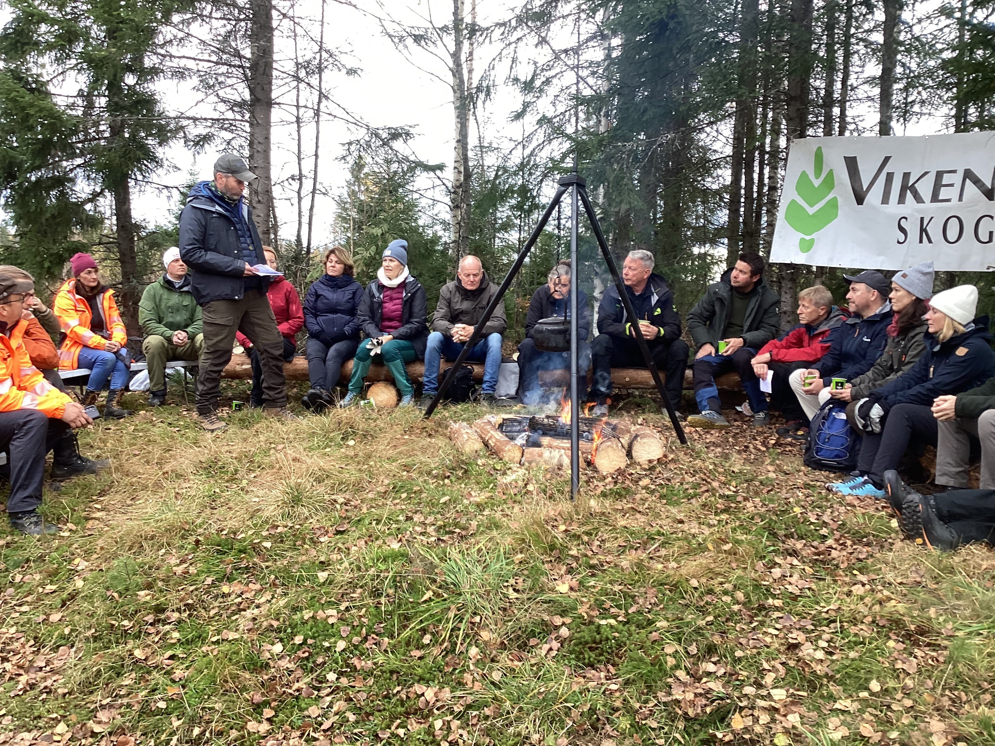 Bilde som viser mennesker som sitter på tømmerstokker rundt et bål, kaffe, skog
