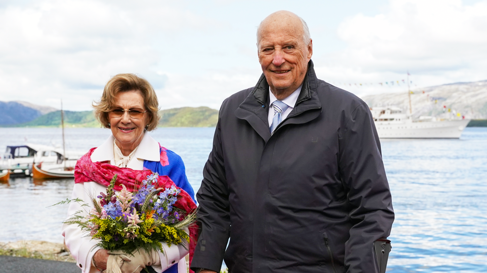 Bildet viser det norske Kongeparet ved havet. Kongeskipet ligger i bakgrrunnen.