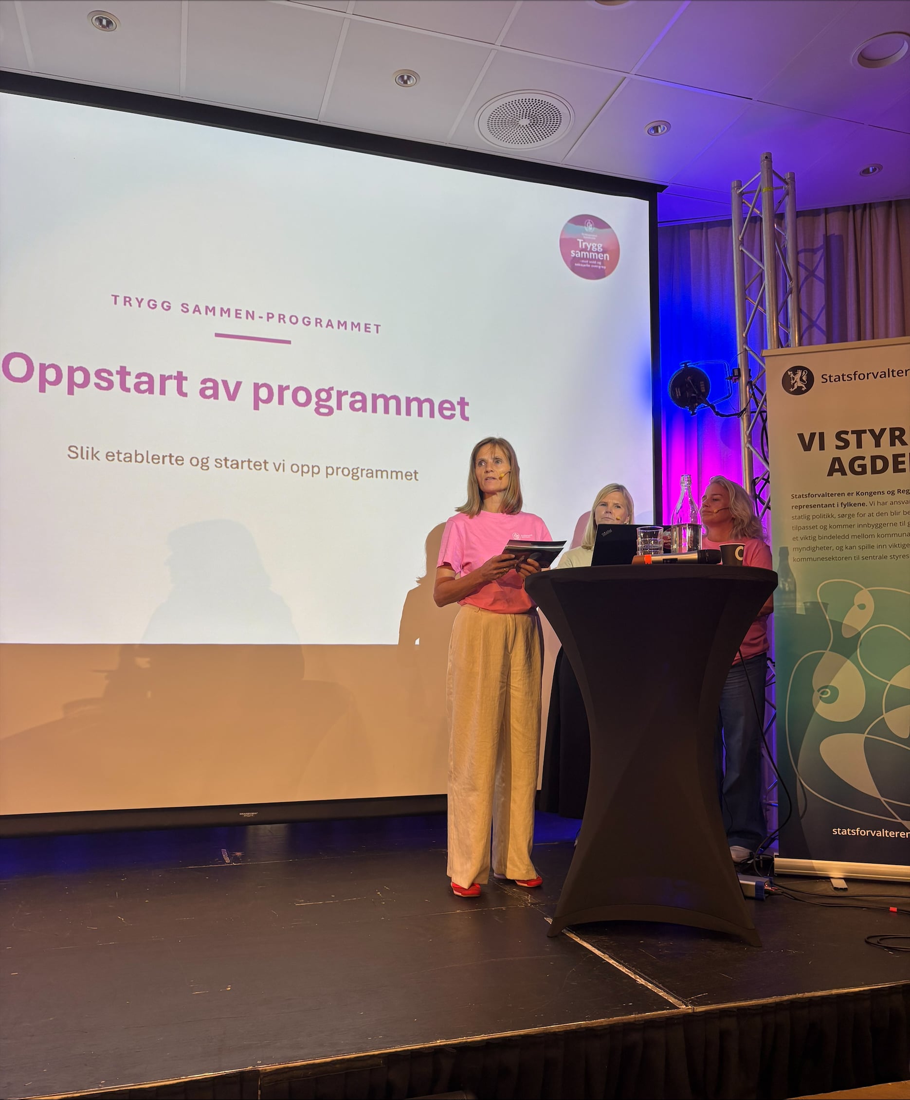 Tre representanter fra Kristiansand kommune står på scenen under et seminar og presenterer Trygg Sammen-programmet. På storskjermen bak vises et lysbilde med tittelen «Oppstart av programmet» og teksten «Slik etablerte og startet vi opp programmet». Innlegget handler om kommunens tverrfaglige arbeid for å forebygge vold og seksuelle overgrep mot barn og unge.