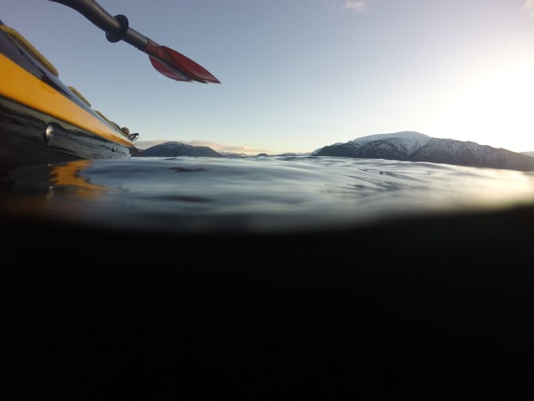 Sjøvatn, svart kajakk, raud padleåre, fjell med snø på toppane