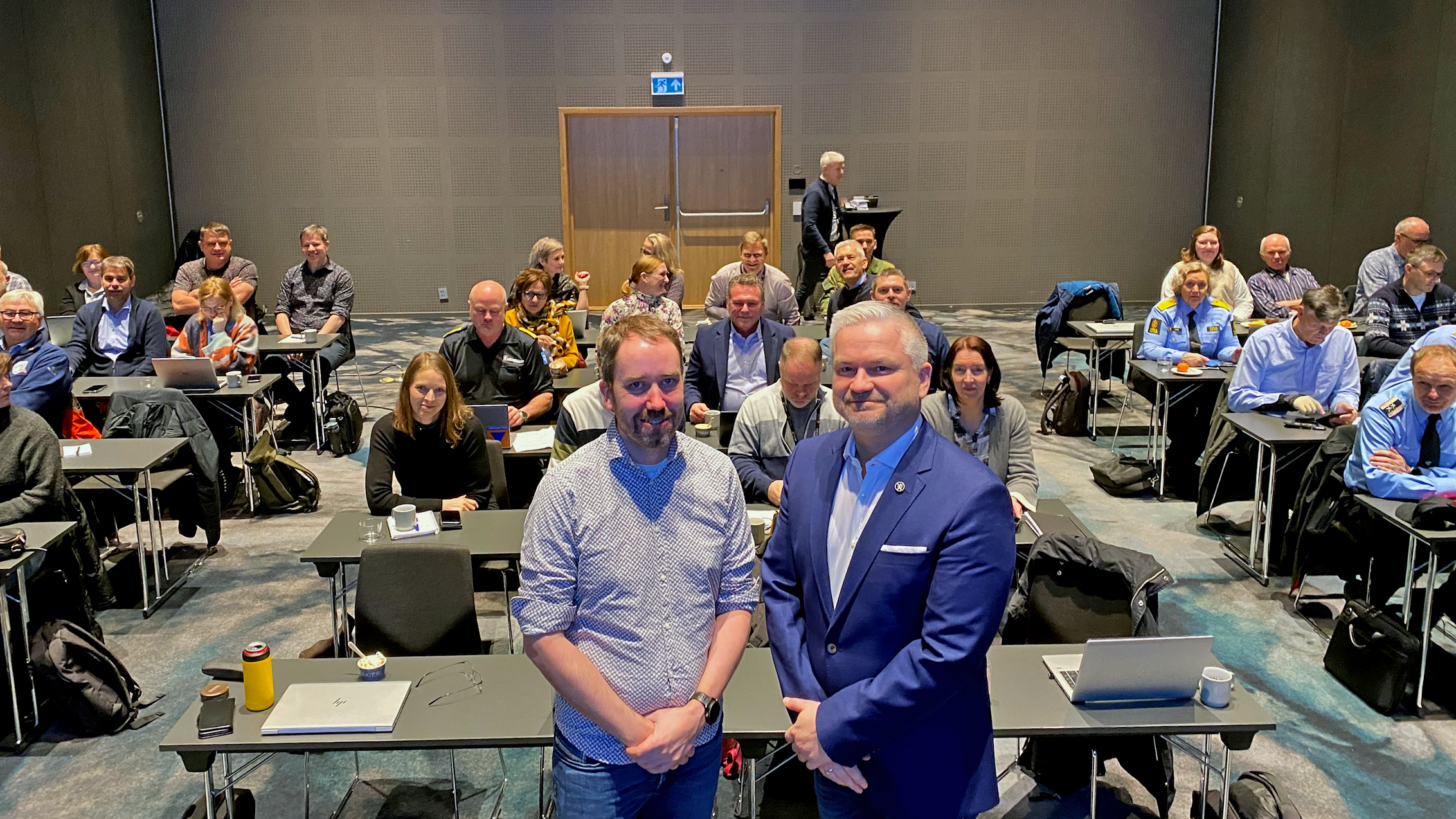 To menn oppstilt foran andre mennesker i konferansesal.