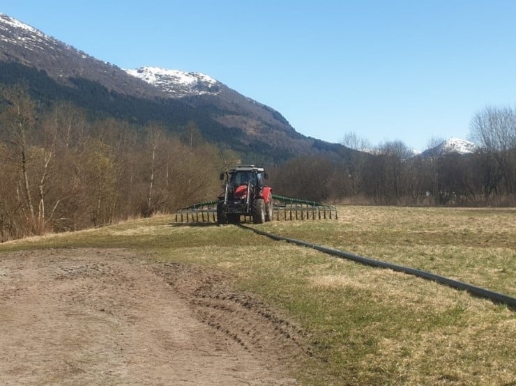 Blå himmel, fjell med snø på toppane, skog, rød traktor, nedlegging av gjødsel med nedleggingsutstyr