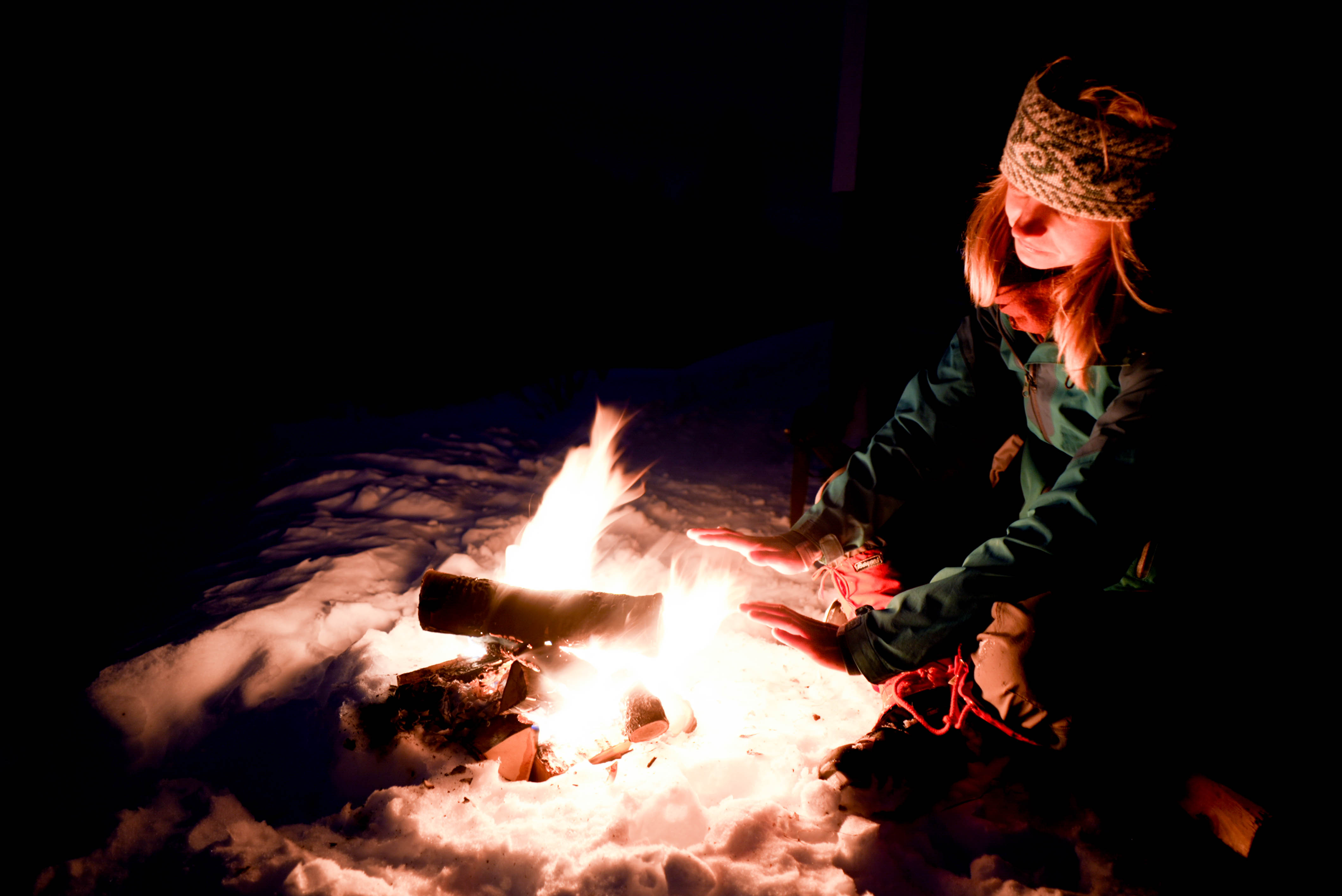En person sitter på snø foran et bål og varmer hendene.