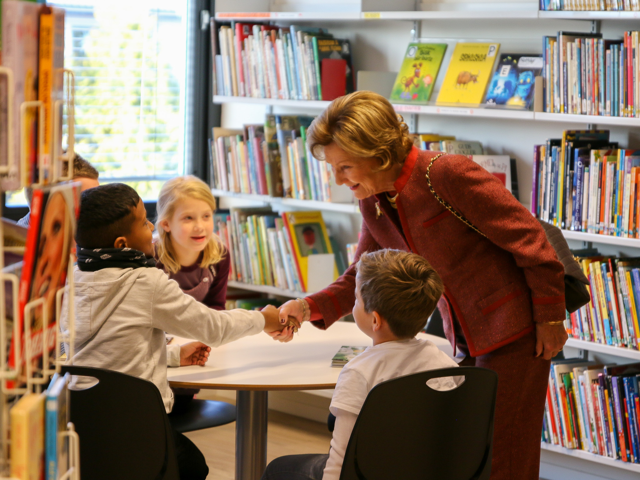 Dronning Sonja hilser på skolebarn i eit bibliotek. 