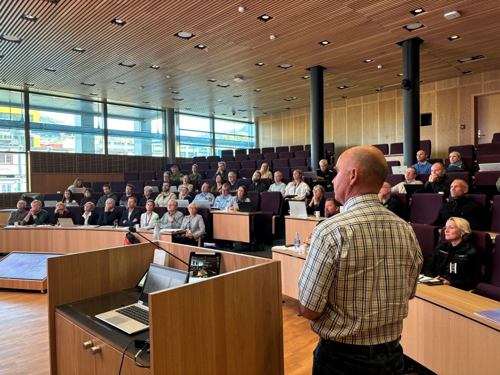 Mann ved talerstol. Mennesker i auditorium.