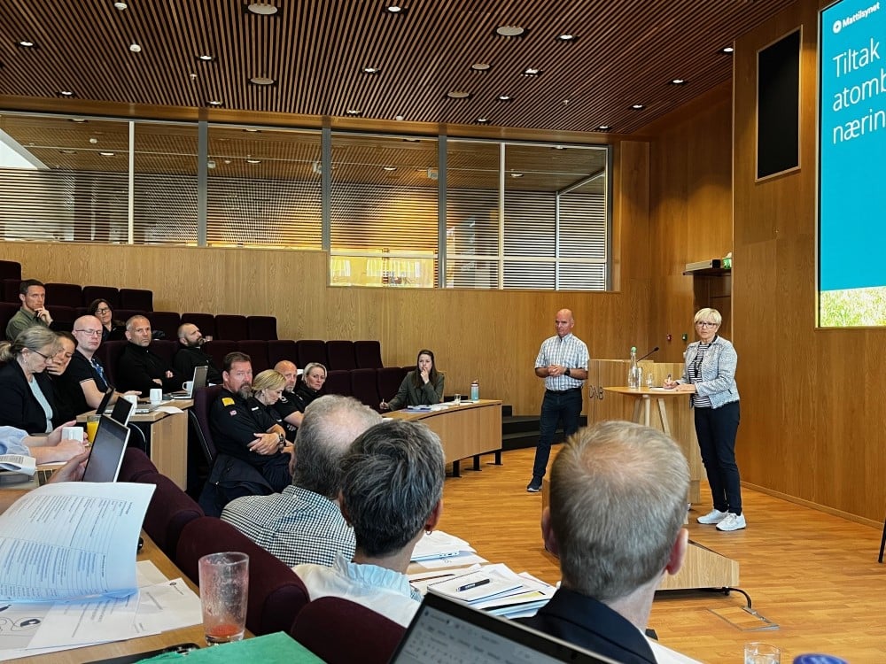 Personar ved talarstol. Personar i auditorium i bakgrunn.