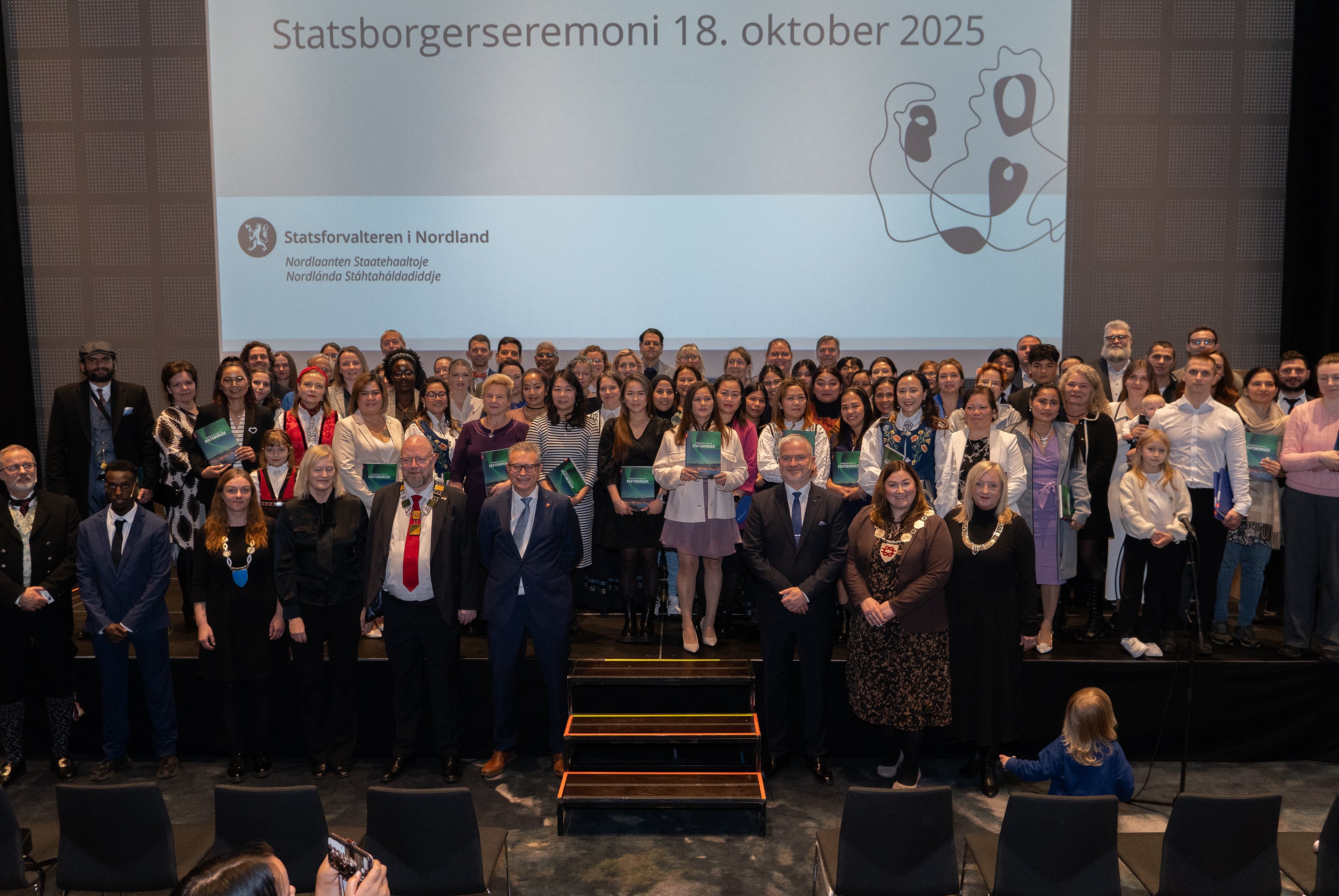 En stor gruppe pent kledde mennesker står oppstilt på en scene.