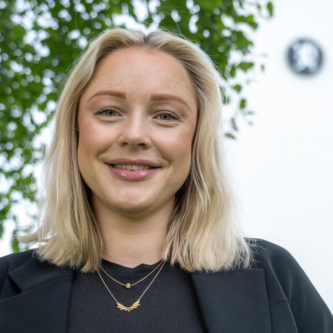 Smilende kvinne med lyst hås og sort dressjakke. Bildet er tatt utendørs foran et tre. Man kan se løvet bak hodet hennes. 