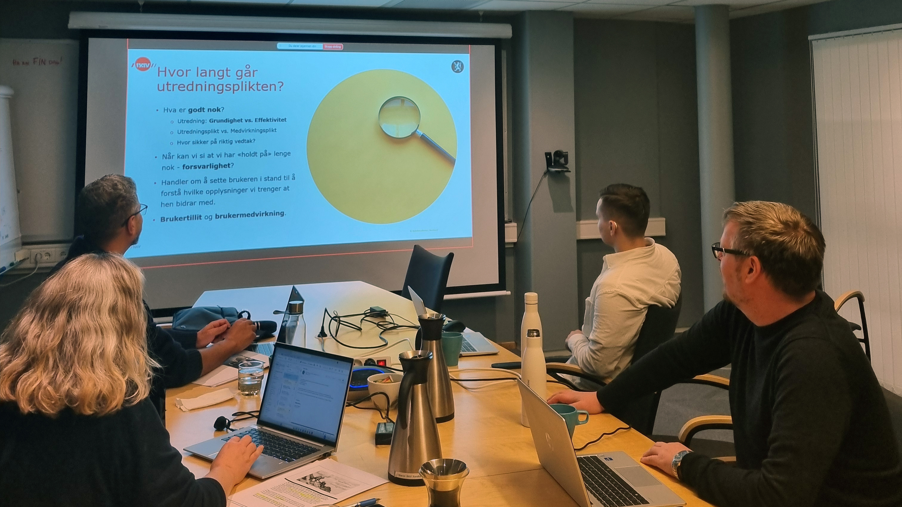 Fire personer sitter rundt møtebord og ser på et Powerpoint-bilde med tekst på et lerret.