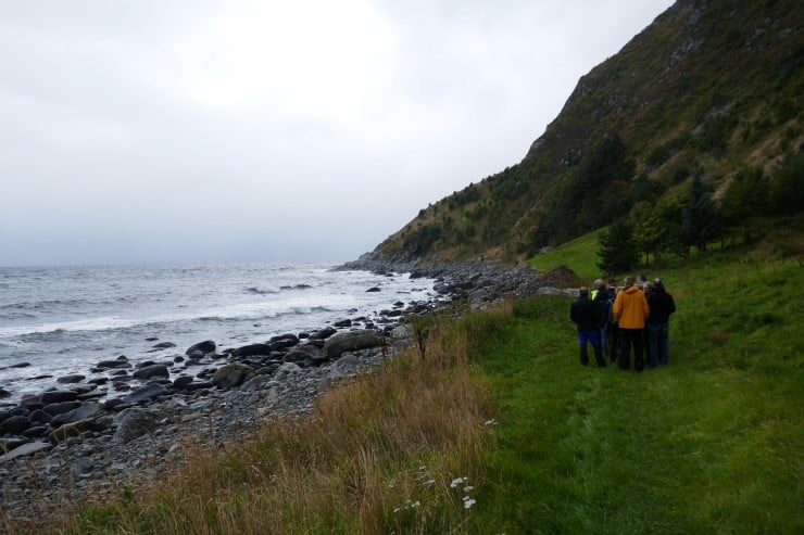 folk som går i flokk på grøn eng langs strand med litt gråvær og litt bølgjer