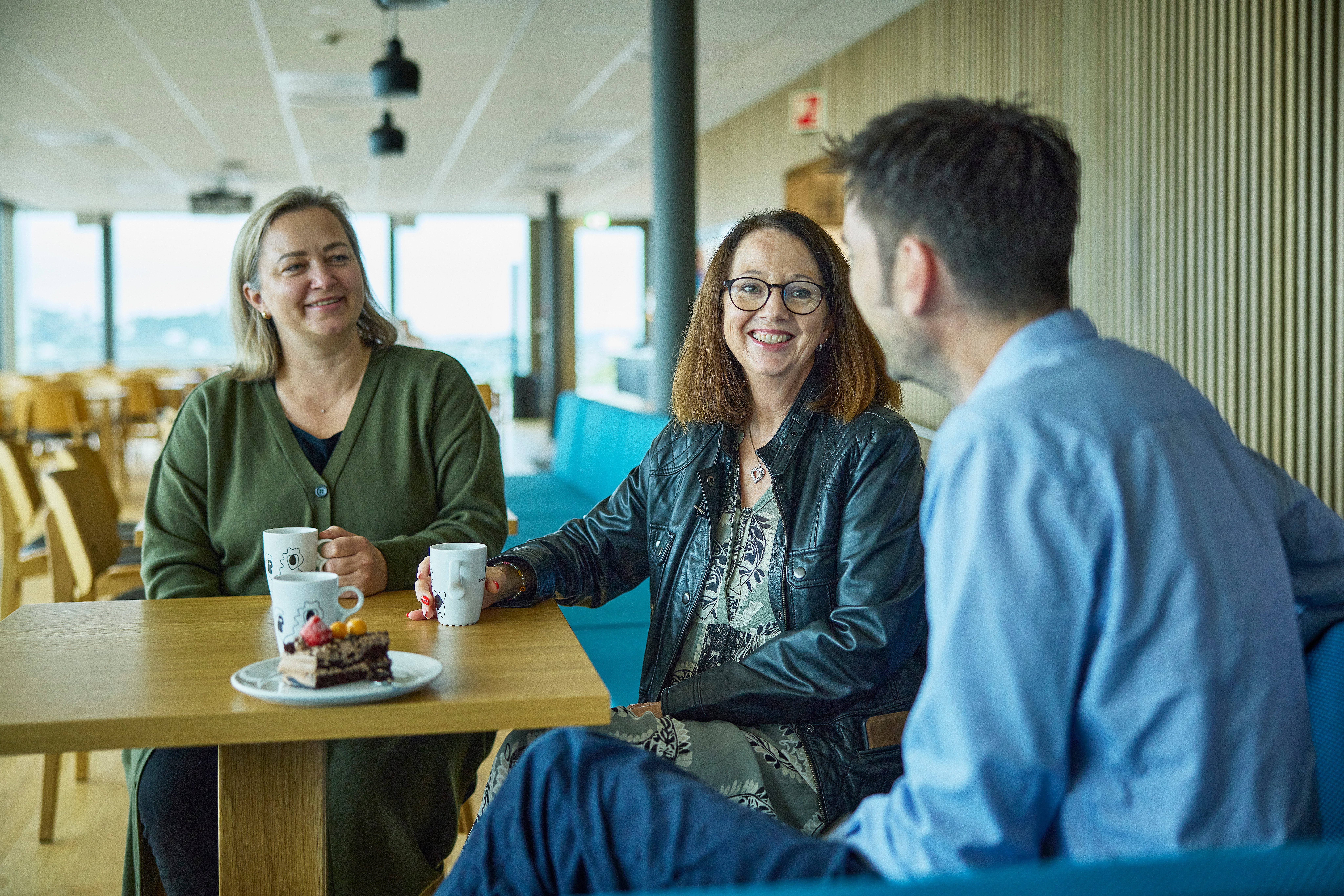 To kvinner og én mann sitter rundt bord i kantinen, hvor de drikker kaffe og spiser kake