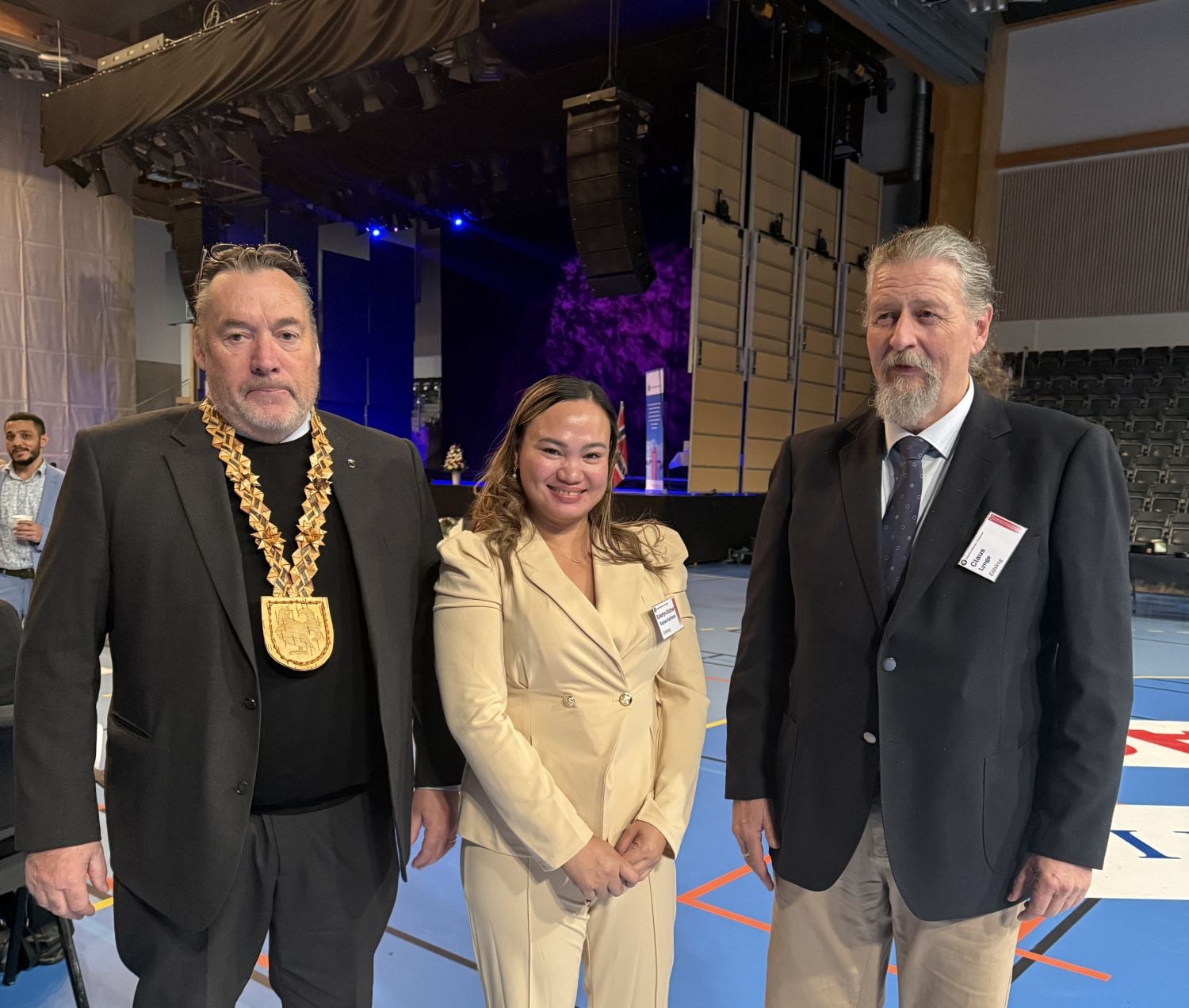 En kvinne og to menn fra eidskog står oppstilt i en festpyntet hall. 