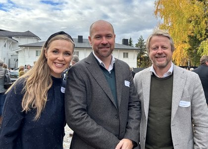 Tre mennesker, ei kvinne og to menn, ved siden av hverandre. Bildet er tatt utendørs. I bakgrunnen skimtes et hvitt hus og noen trær i høstfarger.