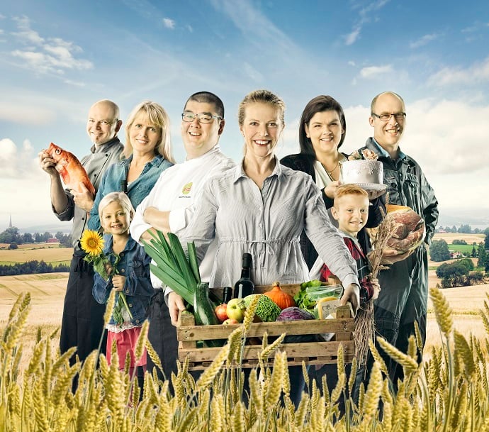 Bildet viser åtte smilende mennesker av ulikt kjønn og alder som står ute i en moden kornåker. De holder ulike matprodukter, blomster eller kornaks i hendene. I front står ei blid, lyshåra kvinne med ei trekasse full av grønsaker.