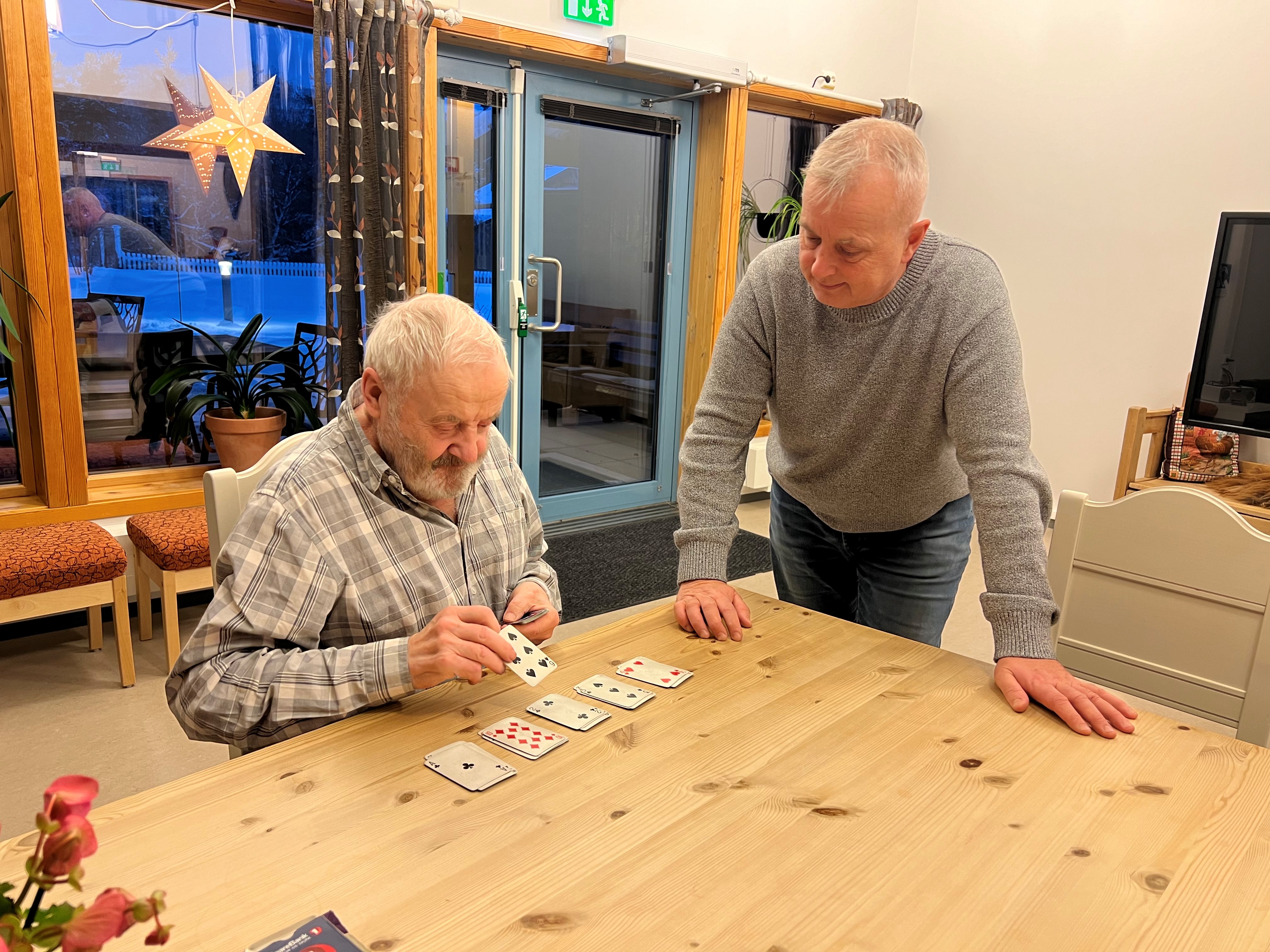 To eldre menn snakker sammen ved et bord. Den ene legger kabal.